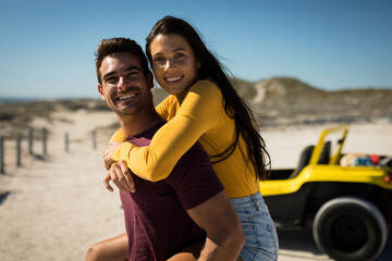Happy caucasian couple next to beach buggy by the sea piggybacking