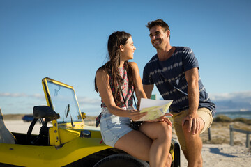 Happy caucasian couple sitting on beach buggy by the sea reading roadmap smiling