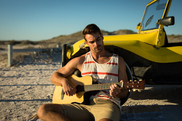 Happy caucasian man leaning against beach buggy by the sea playing guitar