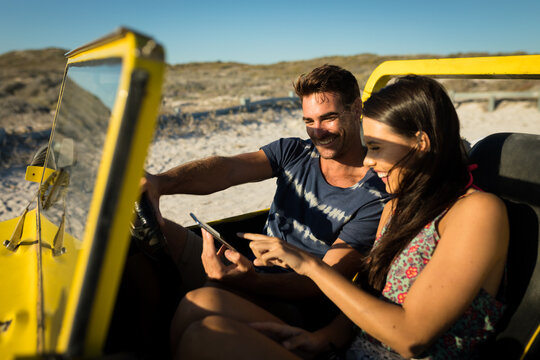 Happy caucasian couple sitting in beach buggy by the sea using smartphone