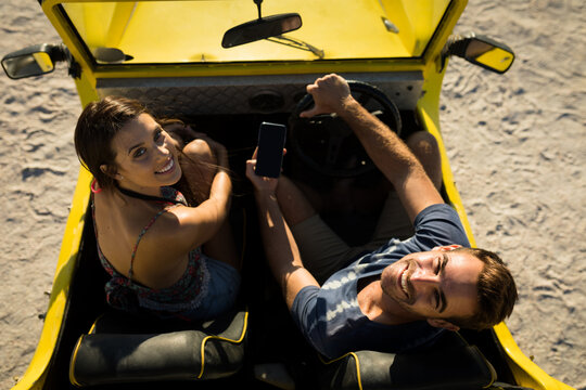 Happy caucasian couple sitting in beach buggy by the sea using smartphone