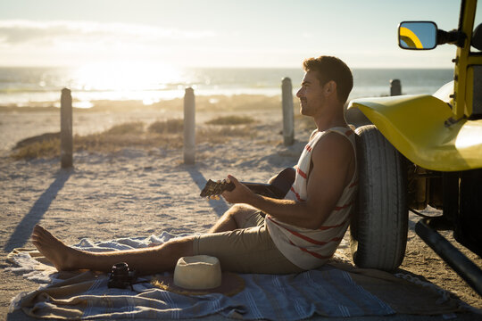 Happy caucasian man leaning against beach buggy by the sea playing guitar - Powered by Adobe