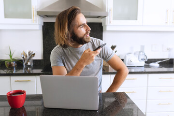Caucasian man sitting in kitchen using laptop and talking on smartphone