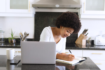 Fototapeta na wymiar African american woman sitting in kitchen using laptop and taking notes