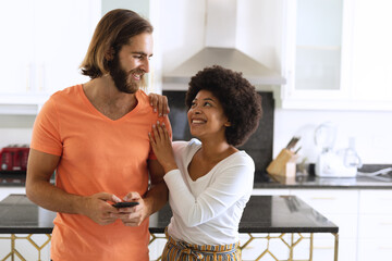 Happy diverse couple in kitchen using smartphone and embracing