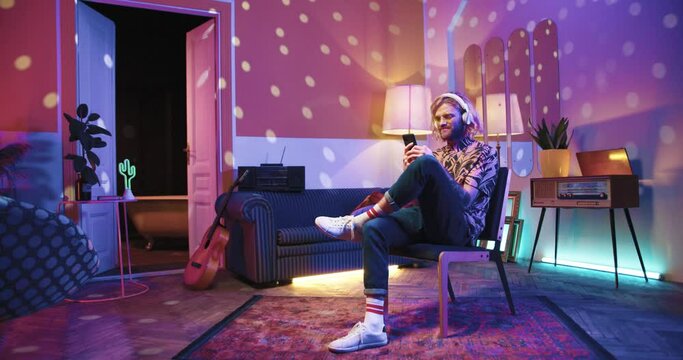 Handsome young Caucasian stylish man sitting on chair listening to music in headphones enjoying fav song while resting at home in living room with warm light and texting on smartphone browsing online