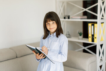 Cheerful teen girl at casual clothes studies at home. Girl makes some notes while standing at the sofa in living room.