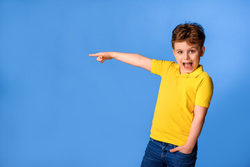 Happy little boy kid in the yellow t-shirt is pointing with his hand and laughing on blue background.