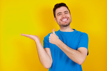 young handsome caucasian man wearing blue t-shirt against yellow background Showing palm hand and doing ok gesture with thumbs up, smiling happy and cheerful.