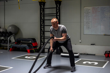 Fototapeta na wymiar Strong young man working out with battle ropes in a cross training gym. Shot of a man training with heavy ropes at the gym. Determined guy using battle rope while doing physical training.