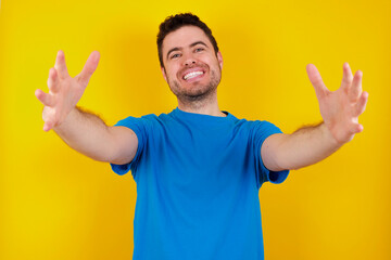 young handsome caucasian man wearing blue t-shirt against yellow background looking at the camera smiling with open arms for hug. Cheerful expression embracing happiness.