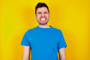 Positive young handsome caucasian man wearing blue t-shirt against yellow background smiles happily, glad to receive pleasant news from interlocutor, keeps palms together, People emotions concept.