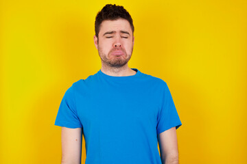 Fototapeta premium young handsome caucasian man wearing blue t-shirt against yellow background keeps teeth clenched, frowns face in dissatisfaction, irritated because of much duties.
