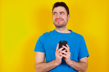 Shocked young handsome caucasian man wearing blue t-shirt against yellow background opens mouth hold phone reading advert unbelievable big shopping prices
