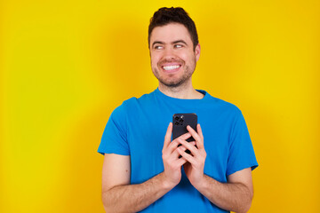 young handsome caucasian man wearing blue t-shirt against yellow background holds telephone hands reads good youth news look empty space advert