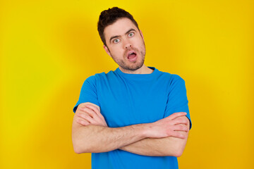 Charming thoughtful young handsome caucasian man wearing blue t-shirt against yellow background stands with arms folded concentrated somewhere with pensive expression thinks what to do