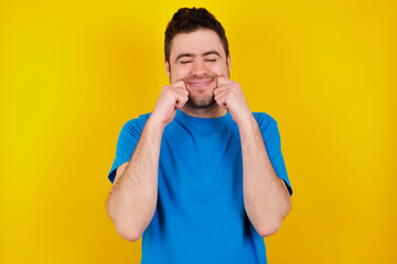 Pleased young handsome caucasian man wearing blue t-shirt against yellow background with closed...