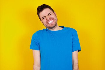 Obraz premium Positive young handsome caucasian man wearing blue t-shirt against yellow background with overjoyed expression closes eyes and laughs shows white perfect teeth