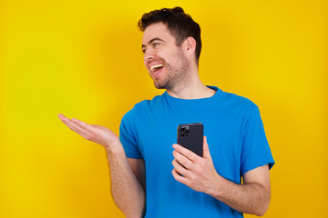 Happy pleased young handsome caucasian man wearing blue t-shirt against yellow background raises palm and holds cellphone uses high speed internet for text messaging or video calls