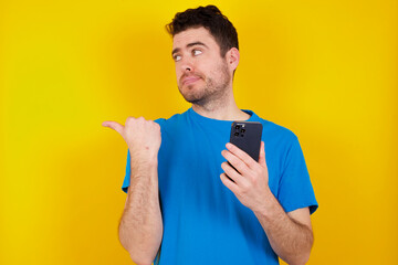 young handsome caucasian man wearing blue t-shirt against yellow background points thumb away and shows blank space aside, holds mobile phone for sending text messages.