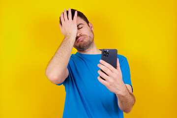 Upset depressed young handsome caucasian man wearing blue t-shirt against yellow background makes face palm as forgot about something important holds mobile phone expresses sorrow and regret blames