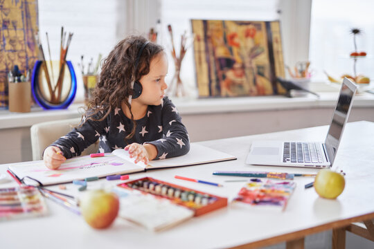 Cute small kid girl in handsfree headphones sitting at a table using a laptop computer for education or homework doing. Doing business or small businesswoman concept. High quality photo