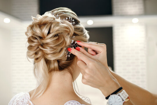 Preparation for the wedding of the bride in the salon. A master hairdresser with a red manicure straightens the bride's hair by inserting a brooch. Rear view of a happy bride in a white dress