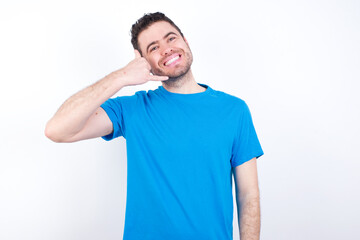 Obraz premium young handsome caucasian man wearing blue t-shirt against white background smiling doing phone gesture with hand and fingers like talking on the telephone. Communicating concepts.