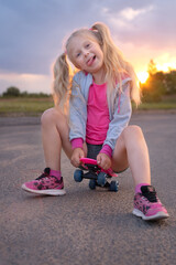 Cute little blonde girl sitting on the pink skating board and posing with funny face