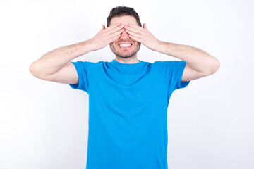 young handsome caucasian man wearing blue t-shirt against white background covering eyes with hands smiling cheerful and funny. Blind concept.