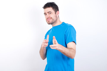 Joyful young handsome caucasian man wearing blue t-shirt against white background wink and points...