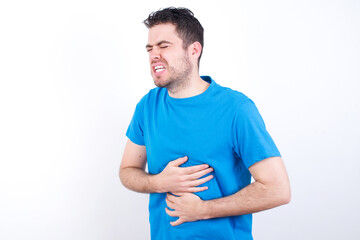 Obraz premium young handsome caucasian man wearing blue t-shirt against white background with hand on stomach because nausea, painful disease feeling unwell. Ache concept.