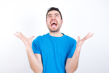 Fototapeta premium young handsome caucasian man wearing blue t-shirt against white background crazy and mad shouting and yelling with aggressive expression and arms raised. Frustration concept.