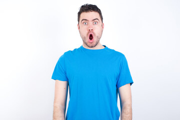 Oh my God. Surprised young handsome caucasian man wearing blue t-shirt against white background stares at camera with shocked expression exclaims with unexpectedness,