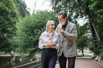 Fototapeta premium Full length portrait of happy senior couple walking and talking at green park. Bearded husband gently hugging beautiful wife. Pensioners enjoying time together.