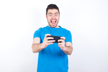 Crazy impressed young handsome caucasian man wearing blue t-shirt against white background use cell phone reads incredible news on social media information stare, screams wow omg.