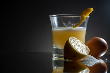 Whisky sour with egg white and lemon zest on the top of the glass. Single egg, half of lemon and sugar clod on the side. Reflecting surface and dark gradient background