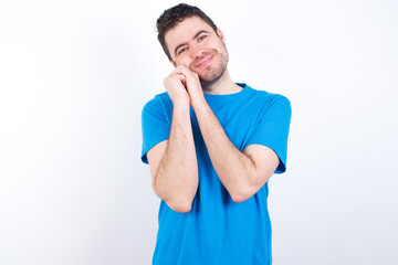 Charming serious young handsome caucasian man wearing white t-shirt against white background keeps hands near face smiles tenderly at camera