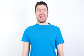 Fototapeta premium Funny young handsome caucasian man wearing white t-shirt against white background makes grimace and crosses eyes plays fool has fun alone sticks out tongue.