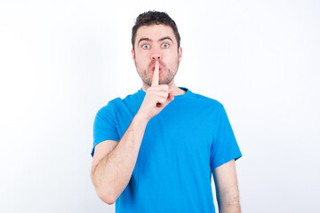 Surprised young handsome caucasian man wearing white t-shirt against white background makes silence gesture, keeps finger over lips and looks mysterious at camera