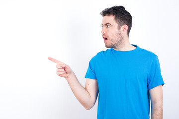 Stunned young handsome caucasian man wearing white t-shirt against white background with greatly surprised expression points away on copy space, indicates something