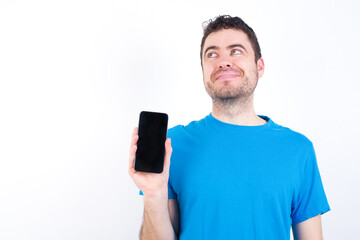 young handsome caucasian man wearing white t-shirt against white background holds new mobile phone and looks mysterious aside shows blank display of modern cellular