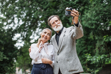 Beautiful senior family in stylish clothes taking self portrait on retro camera. Happy wife and husband standing at green park and enjoying sunny weather.