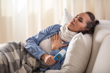 Fototapeta premium Ill woman experiencing breathing and cough problems while holding forehhead thermometer and lying down on a sofa