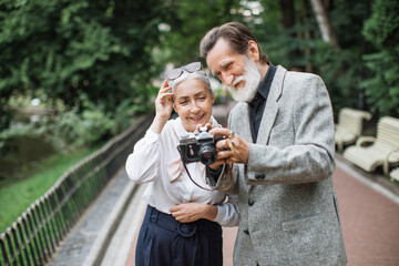Smiling aged man and woman standing at green park and viewing photos on retro camera. Lovely mature couple walking outdoors and taking pictures.
