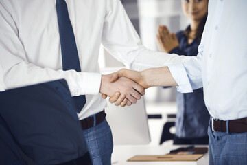 Obraz premium Two businessmen are shaking hands in office, close-up. Happy and excited business woman stands with raising hands at the background. Business people concept