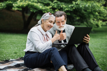 Happy mature couple using modern laptop for video call on fresh air. Stylish senior man and woman sitting on grass, smiling and showing thumbs up.