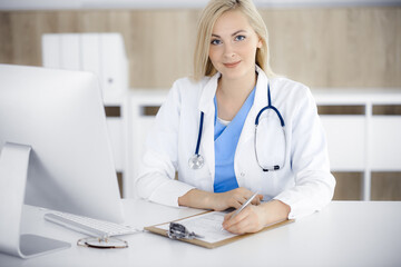 Portrate of woman-doctor at work while sitting at the desk in clinic. Blonde cheerful physician filling up medical form or prescription