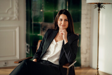 Portrait of a business woman brunette is in a modern stylish office. A satisfied manager is a finance lawyer. Sitting on a chair thinking about work. Clothing business suit in black.