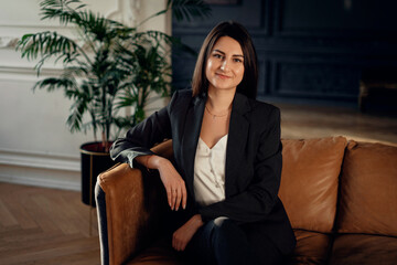 Sitting on a leather sofa, thinking about work. Portrait lawyer business woman brunette of European appearance is located in a modern stylish office coworking. A confident financial psychologist.